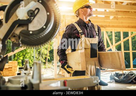A worker carrying wooden planks stands at a construction site during daylight. The man wears safety gear and sunglasses. Tools are visible in the back Stock Photo