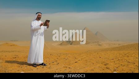 Confident Emirati businessman in white kandura using a digital tablet standing in the desert before the Giza pyramids in Egypt ideal for MENA Stock Photo