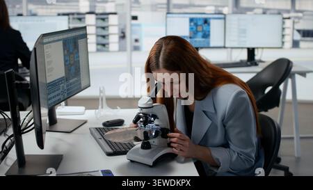 Woman using microscope in office, reviewing semiconductor substrate structures. Female materials scientists analyzing silicon wafer with optical device in industrial plant RD workspace, camera A Stock Photo
