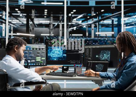Engineer using a wheelchair works with a colleague to configure AI and machine learning equipment in an inclusive server room. Stock Photo