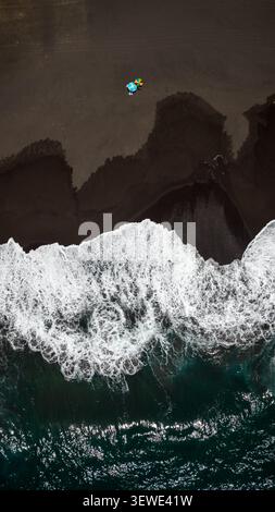 Aerial view of colorful beach umbrellas on the black sand of Ribeira Grande beach with crashing white waves meeting the dark volcanic sand in Ribeira Stock Photo
