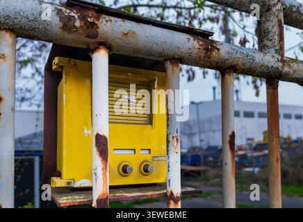 Old yellow industrial intercom mounted behind a weathered rusty metal fence at the entrance of a commercial or industrial site. Stock Photo