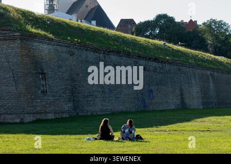 Two people relaxing on a lush green lawn next to a historic brick fortress wall under bright sunlight, Europe. Stock Photo