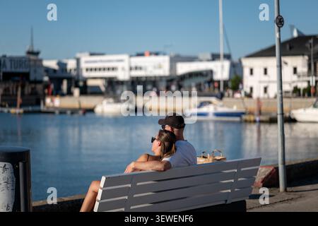 Romantic couple sitting on a white wooden bench at the harbor, looking out at the sea, boats, and marina on a sunny day. Stock Photo