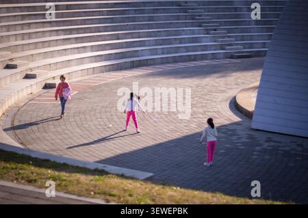 Children playing badminton in a spacious concrete amphitheater plaza with long afternoon shadows stretching across the ground. Stock Photo
