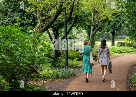 Two women walking together along a tree-lined dirt path in a peaceful lush park. Stock Photo