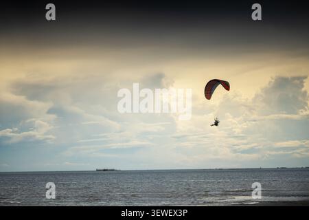 A person enjoying an adventurous flight while paragliding over the open ocean during a cloudy sunset, showcasing outdoor recreation and scenic natural Stock Photo