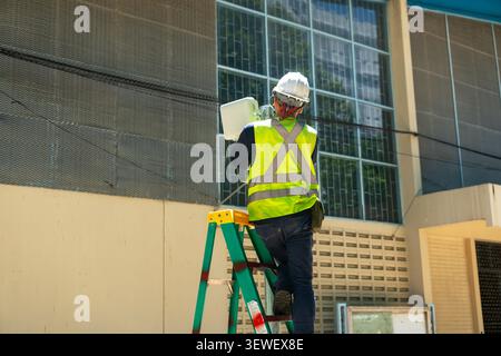 A construction worker wearing safety gear and helmet is standing on a ladder near a building with glass windows, actively engaged in a building or mai Stock Photo
