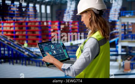 Mature Woman Uses Laptop To Monitor Warehouse Inventory And Delivery Data. Stock Photo