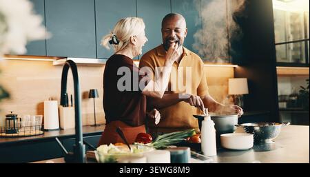 Cooking, laughing and taste with interracial couple in kitchen of home together for meal preparation. Bonding, feeding and having fun with mature Stock Photo
