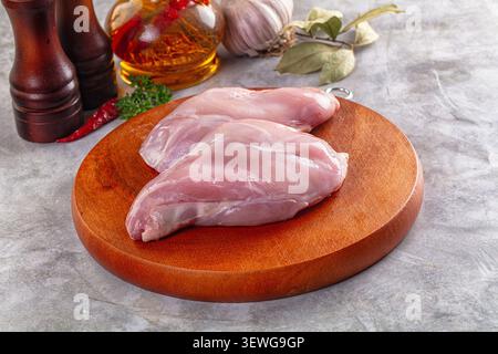 Raw rabbit thighs on round wooden board. Background includes pepper mill, chili, garlic, bay leaves, oil bottle. Soft lighting, neutral gray surface. Stock Photo