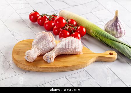 Raw chicken drumsticks on wooden cutting board surrounded by cherry tomatoes leek garlic. Bright red tomatoes contrast with pale chicken green leek. C Stock Photo