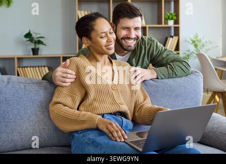 Smiling couple paying online with laptop on sofa at home, browsing shopping apps Stock Photo