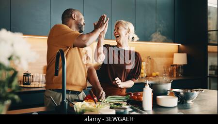 Cooking, high five and laughing with interracial couple in kitchen of home together for meal preparation. Bonding, having fun and success with mature Stock Photo