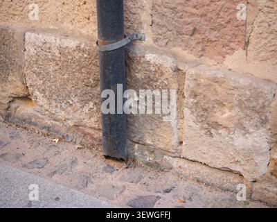 Old, rough hewn stone wall featuring a dark, rusty downspout and part of a cobblestone pavement Stock Photo