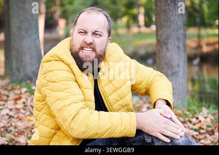 Calm explorer seated beside water body. Tranquil individual with beard waits amid colorful fallen leaves. Relaxed man sporting beard and yellow jacket sitting peacefully in autumn park setting Stock Photo