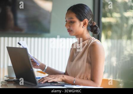 Businesswoman typing on laptop while holding financial documents at office desk, analyzing reports for accounting, budget planning, digital workflow, Stock Photo