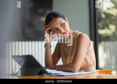 Thoughtful businesswoman looking away while working with tablet and financial papers in office, showing decision making, digital finance, accounting r Stock Photo