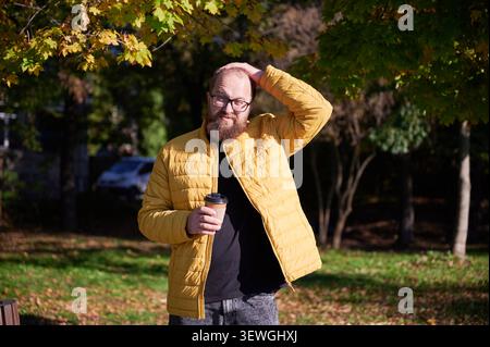 Man with coffee. Reflective man in park. Casual man enjoying autumn morning. Male figure wearing yellow jacket in sunlight. Person with disposable cup exudes calmness during peaceful park morning Stock Photo