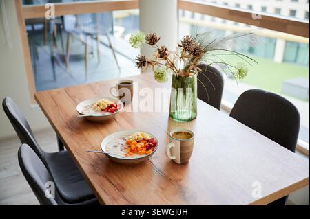 Breakfast served on dining table, bowls of cereal with fruit and cups of tea or coffee in modern home interior, wooden table by window with natural light, concept of healthy morning routine Stock Photo