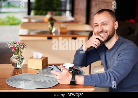 Smiling man holding smartphone at cafe table, resting chin on hand, enjoying coffee break, looking at camera, relaxed mood, modern lifestyle and connection concept Stock Photo