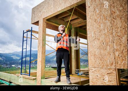 Worker in hardhat assessing measurements of house framework, representing design implementation, accuracy control, construction planning and site operations Stock Photo