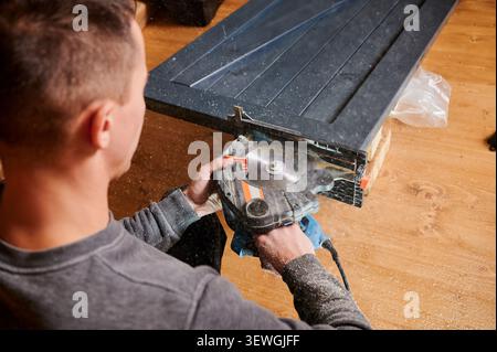 Man cutting wooden panel using circular saw, holding tool firmly while trimming edge. Close up of woodworking process with flying sawdust, renovation and carpentry concept Stock Photo