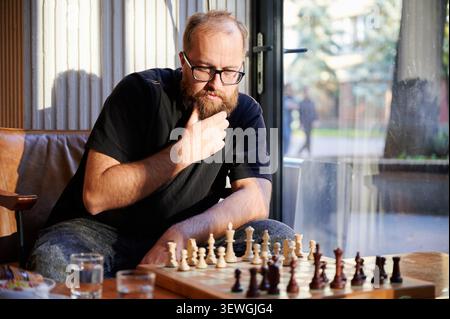 Bearded man studying chessboard under sunlight, hand on chin, focused expression, casual clothing, wooden board displaying midgame, cup and pastry on table, cozy cafe interior, soft window reflections Stock Photo