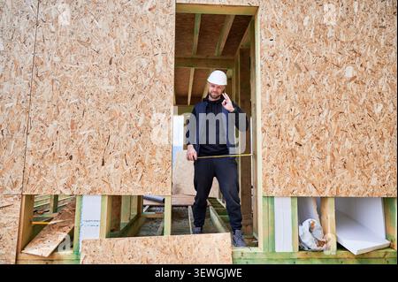 Construction worker standing in doorway of wooden frame house, holding tape measure and showing OK gesture to confirm correct dimensions and successful work Stock Photo