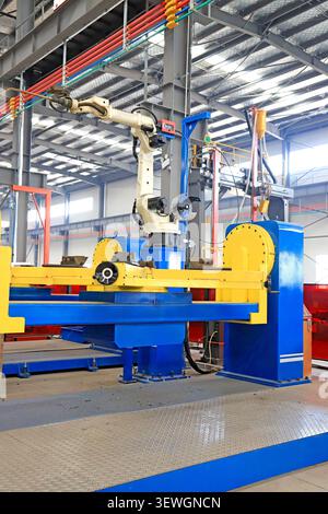 A high-resolution, wide-angle shot of an automated welding robot arm mounted on a vibrant blue and yellow heavy-duty machine in a spacious factory wor Stock Photo
