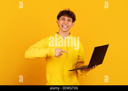 Smiling guy in yellow hoodie holding open laptop and pointing to screen Stock Photo