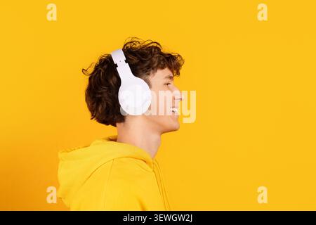 Side profile of smiling young guy in yellow hoodie wearing white headphones Stock Photo