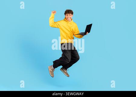 Excited young guy in yellow hoodie jumping with laptop on blue background Stock Photo