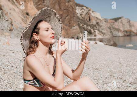 Woman makeup beach, young lady applying lipstick looking at smartphone mirror while sitting on pebble shore in bright sunny summer day Stock Photo