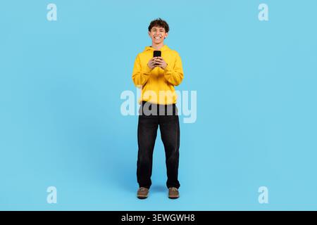 Smiling young guy in yellow hoodie standing with smartphone on blue background Stock Photo