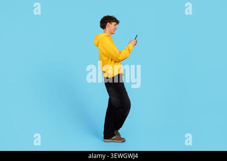 Young guy in yellow hoodie using smartphone on blue studio background Stock Photo