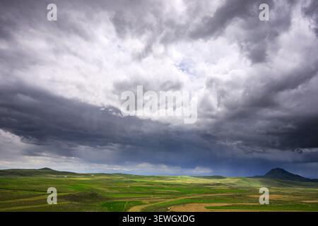 Dark storm clouds gather over green rolling hills. Dramatic sky looms above vast open field. Landscape features grassy plains and distant hills Stock Photo