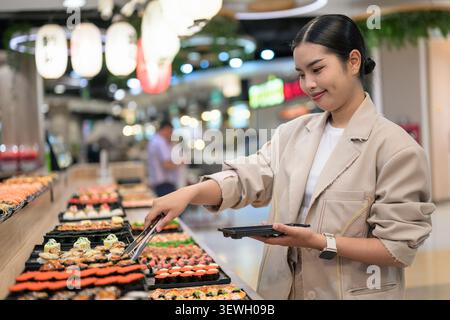A happy young Southeast Asian Thai businesswoman from the Isan region smiling while using tongs to choose food at a vibrant sushi buffet line. She is Stock Photo