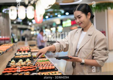 A happy young Southeast Asian Thai businesswoman from the Isan region smiling while using tongs to choose food at a vibrant sushi buffet line. She is Stock Photo