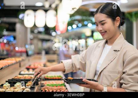 A happy young Southeast Asian Thai businesswoman from the Isan region smiling as she uses tongs to place a piece of sushi onto her takeaway tray. She Stock Photo