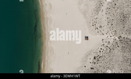 Aerial view of turquoise ocean waters meeting the pristine white sand beach with a small lifeguard station and scattered blue umbrellas on the coast o Stock Photo