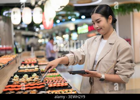 A happy young Southeast Asian Thai businesswoman from the Isan region smiling while using tongs to choose food at a vibrant sushi buffet line. She is Stock Photo