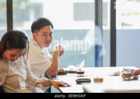 Man smiles gently at table companion. Woman leans forward, focused on plate. Coffee cups and small dishes sit neatly. Natural light streams through la Stock Photo