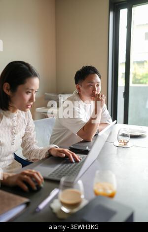Two professionals focus intently on laptop screens. Natural light streams through large glass doors. Soft tones frame a calm, collaborative workspace. Stock Photo