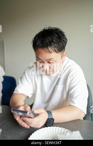 Young man checks phone at desk. Focused gaze, relaxed posture. Casual white shirt, dark watch. Neutral expression, quiet concentration. Ideal for work Stock Photo