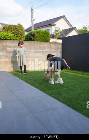 Two girls play on a green artificial turf lawn. One bends down to roll a ball. The other watches with a gentle smile. Modern homes surround the bright Stock Photo