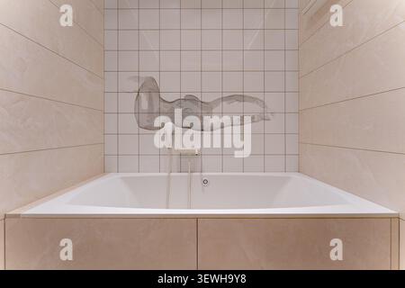 Modern minimalist bathroom interior with built in white bathtub, beige marble tiles and decorative ceramic wall mural in contemporary luxury home Stock Photo