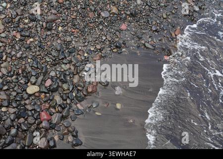 Abstract nature background capturing the transition between wet volcanic sand, multi-colored pebbles, and sea foam. Stock Photo