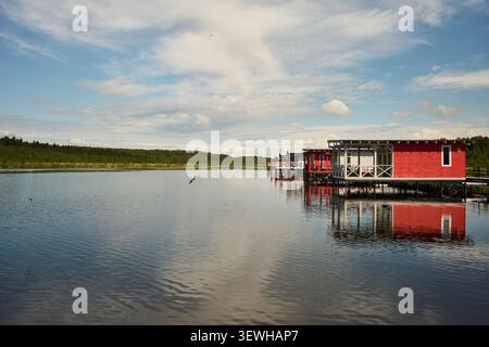 Red cabin on stilts reflected in calm lake at forested shoreline Stock Photo