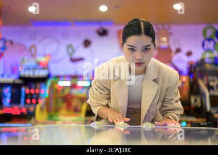 A medium shot taken from across the table showing a focused young Southeast Asian Thai businesswoman from the Isan region leaning over a glowing arcad Stock Photo